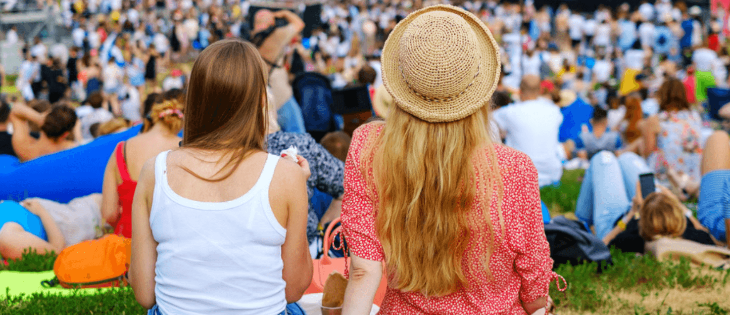 2 women say with their backs to the camera looking over a festival scene, everyone is sat in groups on a field